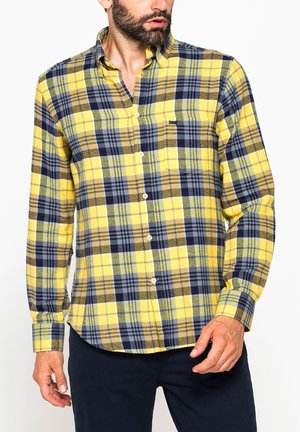 Uomo che indossa una camicia a quadri gialla, blu navy e oliva a maniche lunghe con taschino, abbinata a pantaloni scuri, guardando di lato.