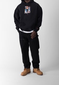 Felpa nera con cappuccio e design grafico, indossata sopra una maglietta bianca. Pantaloni neri a gamba larga e stivali beige con lacci completano l'outfit.