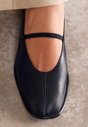 Zapato plano de cuero negro con puntera redondeada, corte decorativo y una banda elástica en la parte superior del pie. Textura suave y diseño elegante.