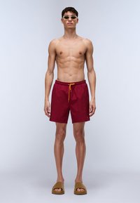 Shorts de bain rouges avec taille élastique et cordon jaune, associés à des sandales à glissière marron. Le modèle est torse nu, portant des lunettes de soleil claires.