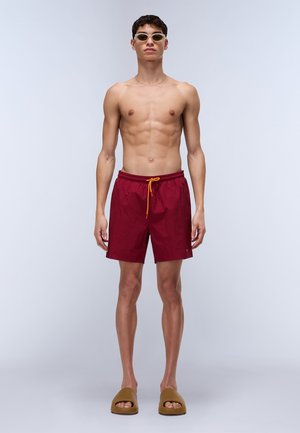 Pantaloni del costume rossi con cintura elastica e cordino giallo, abbinati a sandali scivolabili marroni. Il modello è senza camicia e indossa occhiali da sole chiari.