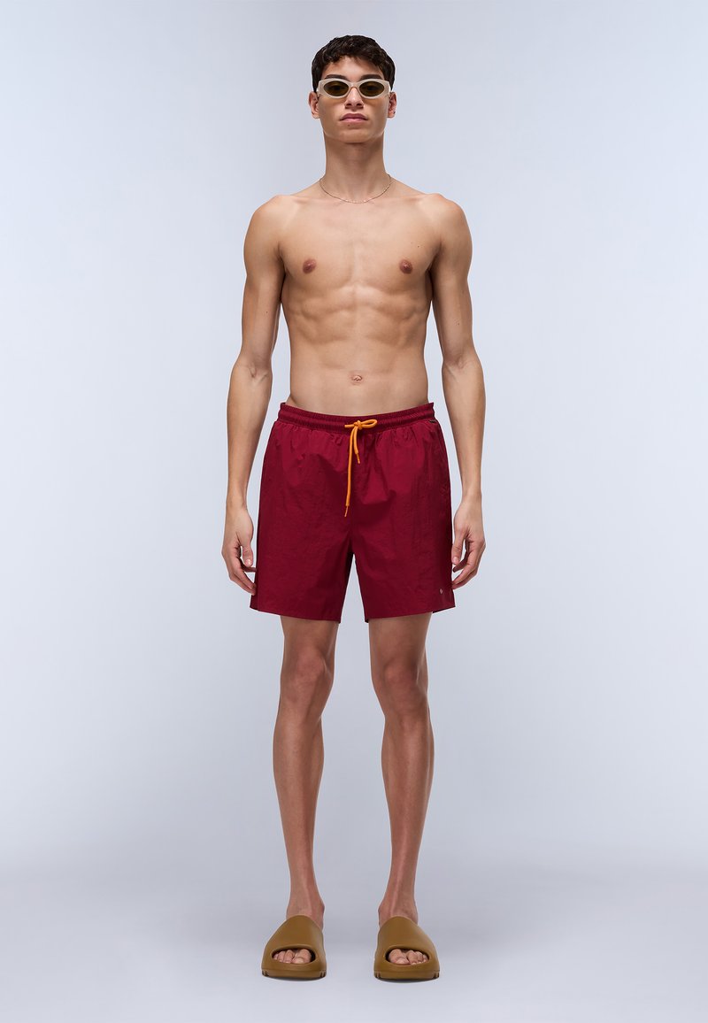 Shorts de bain rouges avec taille élastique et cordon jaune, associés à des sandales à glissière marron. Le modèle est torse nu, portant des lunettes de soleil claires.
