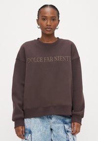 Brūns džemperis ar izšūtu uzrakstu "DOLCE FAR NIENTE" gaiši brūnā krāsā. Ir apaļš kakls un brīvs piegriezums. Kombinēts ar gaiši ziliem kravas stila šortiem.