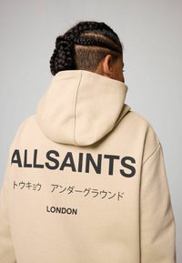 Beżowa bluza z kapturem z czarnym napisem "ALLSAINTS" oraz dodatkowymi azjatyckimi znakami, z dużym kapturem i miękką teksturą.