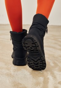 Zwarte geïsoleerde laarzen met een gewatteerd ontwerp, rubberen antislipzolen en zijritsen. Pair gepresenteerd met oranje leggings op een neutrale vloer.