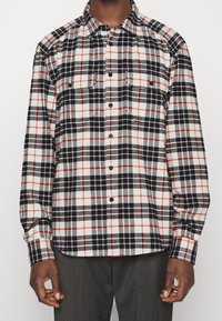 Camicia di flanella a quadri nera, bianca e rossa con maniche lunghe, chiusura a bottoni e due tasche sul petto. Texture morbida e vestibilità rilassata.