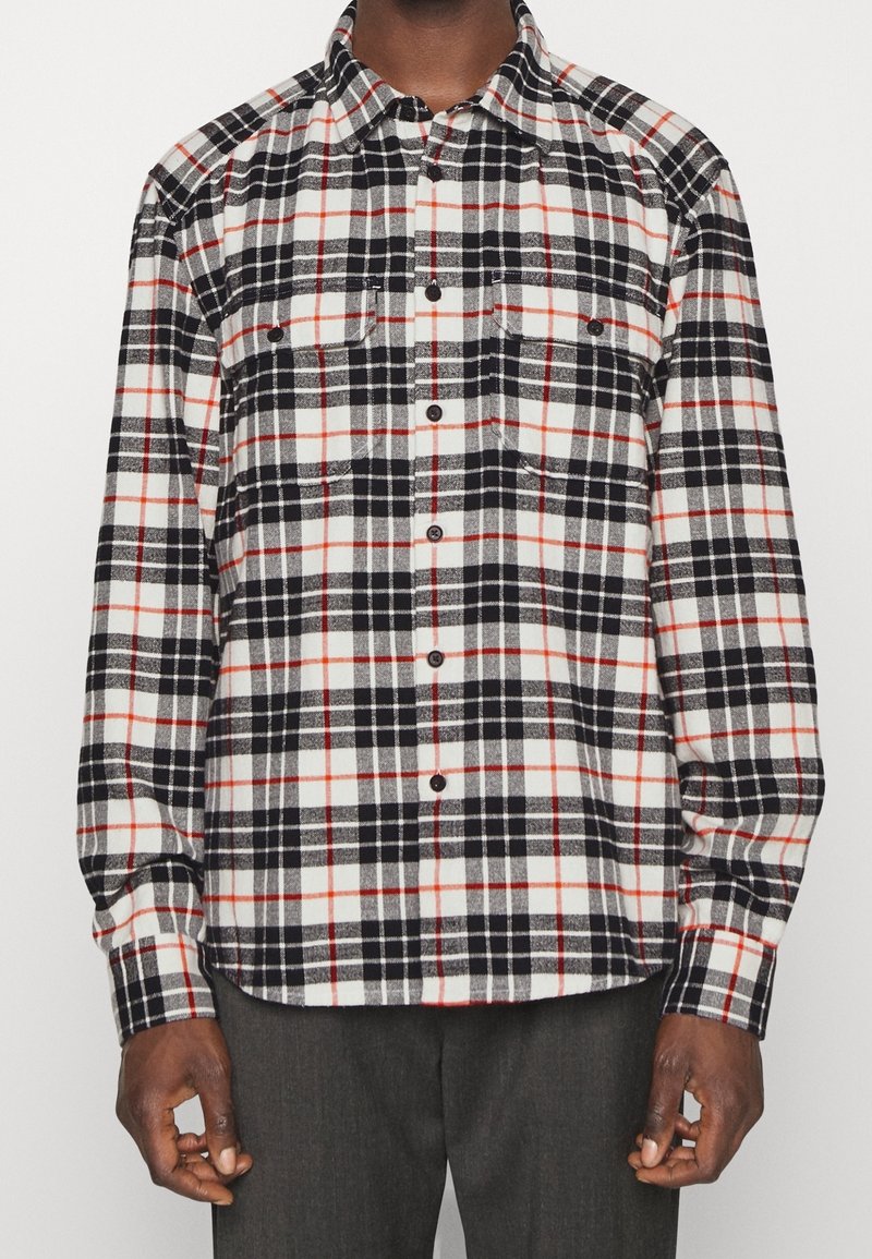 Camicia di flanella a quadri nera, bianca e rossa con maniche lunghe, chiusura a bottoni e due tasche sul petto. Texture morbida e vestibilità rilassata.