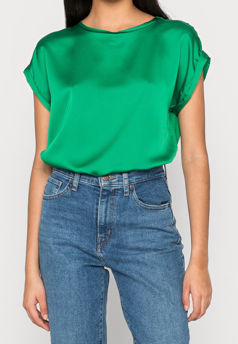 Haut en satin vert avec un col rond et des manches courtes retroussées, associé à un jean denim bleu taille haute. Texture lisse, coupe décontractée.