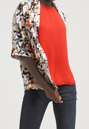 Person iført en knaldrød top under en kortærmet blomsterjakke med mørkeblå jeans, stående mod en ensfarvet lys baggrund.