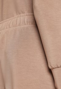 Tissu marron clair avec une texture lisse, présentant une taille élastique et des détails de manches froncées. Vue en gros plan mettant en évidence le matériau.