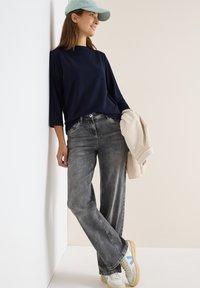 Marineblauw gestructureerd langarmshirt, grijze broek met brede pijpen, houdt een beige geribbeld jack vast, draagt een lichtblauwe pet en crèmekleurige sneakers met accenten.