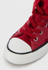 Sapatilha de lona vermelha com biqueira de borracha branca, contorno preto e atacadores vermelhos atados sobre ilhós prateados, sobre um fundo branco.