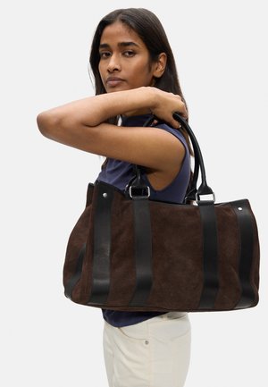 Kobieta w granatowym topie i białych spodniach niesie dużą brązową zamszową i czarną skórzaną paskowaną torebkę na ramieniu.