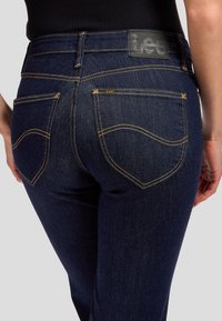 Personne portant un jean Lee bleu foncé avec des coutures jaunes, montrant les poches arrière et un patch logo Lee noir sur la ceinture.