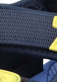 Sandalia de tira de nailon azul que presenta un tejido texturizado, herrajes de acento amarillos y una suela acolchada para mayor comodidad. Primer plano del detalle de la tira.