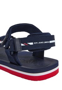 Temno modri sandali z tkani trakovi, ki imajo logotip in bele-rdeče dodatke. Guma z teksturirano površino in večbarvenim dnom.