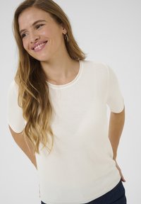 Weißes kurzärmeliges Strickoberteil mit rundem Ausschnitt, weicher Textur und figurbetontem Design. Kombiniert mit dunkelblauer Hose.