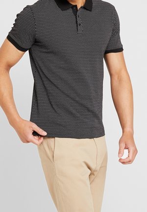 Mand iført sort mønstret poloshirt med beige bukser, holder skørtet på skjorten med venstre hånd.