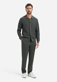 Donkergroene gebreide cardigan met een gestructureerd patroon, lange mouwen en knoopsluiting, gecombineerd met donkergroene broeken en witte sneakers.