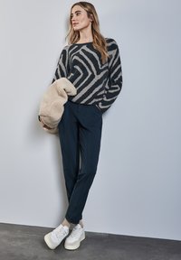 Maglione lavorato a maglia con motivo zigzag nero e beige, abbinato a pantaloni blu e sneakers bianche. Il modello tiene in mano una giacca di peluche beige.