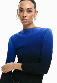 Langärmliges Oberteil mit Farbverlauf von hellem Blau zu dunklem Blau, mit strukturierten Zickzackmustern und rundem Ausschnitt.