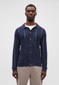 Tengerészkék kötött kardigán gallérral, fából készült gombokkal, texturált mintával, bordázott mandzsettával és szegéllyel. Fehér ing fölé viselve.