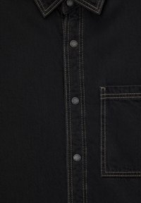 Sort denimskjorte med knappelukning foran, spids krave, kontrasterende beige syninger og en enkelt brystlomme.