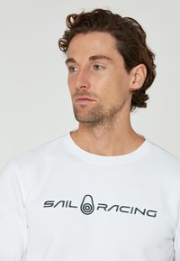 Sudadera blanca con cuello redondo, que presenta un logo bordado en negro de "SAIL RACING" en el pecho. Textura suave y diseño informal.