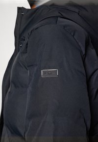 Svart puffjacka med hög krage, texturerat tyg och en märkeslapp på överarmen för identifiering.