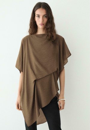 Kvinde med langt bølget hår iført en løs brun asymmetrisk top og sorte bukser, stående foran en ensfarvet lys baggrund.