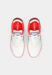 Bela športna obutev z rdečimi poudarki, ki ima perforiran sprednji del, srebrne detajle in teksturiran podplat. Blagovna znamka je na ježku in notranji podplati.