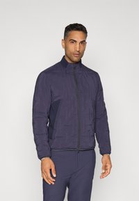 Veste matelassée de couleur marine avec col montant, fermeture éclair et panneaux contrastants. Texture lisse avec des poignets et un ourlet élastiques, fonctionnelle pour le superposition.
