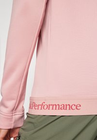 Rožinė sportinė ilgomis rankovėmis palaidinė su lygiu paviršiumi, kurią puošia kontrastingas raudonas "PeakPerformance" logotipas apatinėje ornamente.