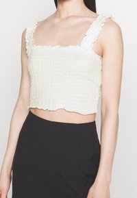Cropped top in light cream with a textured, ribbed pattern. Features ruffled straps and a scalloped hem. Paired with black bottoms.