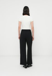 Eine Frau mit schulterlangen schwarzen Haaren, die ein weißes kurzärmliges Oberteil, schwarze weit geschnittene Hosen und schwarze High Heels trägt, steht vor einer weißen Wand.
