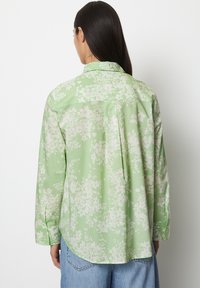 Camicia verde chiaro a maniche lunghe con un motivo floreale bianco. Presenta uno yoke posteriore, orlo curvo e polsini con bottoni. Tessuto morbido.