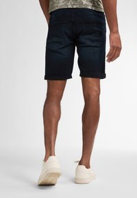 Tummansiniset farkkuhousut, joissa on käärityt lahkeet ja kaksi takataskua, yhdistettyinä beigeihin lenkkitossuihin, esitellään takaa.