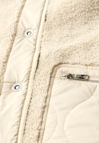 Chaqueta beige con forro de felpa y tejido suave, que presenta un botón a presión y un bolsillo con cremallera en el lateral, destacando un acolchado de costuras.