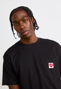 Black cotton t-shirt with short sleeves and a chest pocket. Pocket features a red heart logo with white text. Smooth texture, relaxed fit.