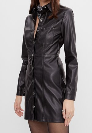 Vrouw die een zwarte nepleren mini-jurk draagt met knopen, lange mouwen en borstzakken, gecombineerd met ringen en een neksjaal.