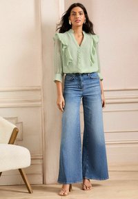 Bluză ușor verde, transparentă, cu accente din volane și buline, asortată cu blugi lejeri din denim deschis. Modelul stă pe un podea din lemn.