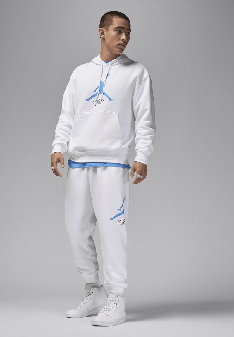 Weißer Kapuzenpullover mit blauem Logo, Fronttasche und Kordelzug. Weiße Jogginghose mit blauen Akzenten und Logo. Kombiniert mit weißen Turnschuhen.