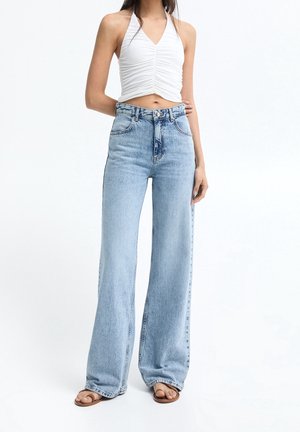 Femme portant un haut à nouer blanc froncé, un jean bleu clair taille haute à jambes larges, et des sandales marron, debout sur fond blanc.