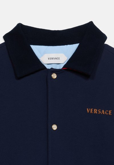 Μαρινέρ μπουφάν Versace με ριμπ λαιμόκοψη, κουμπιά που μοιάζουν με πέρλες, ανοιχτό μπλε εσωτερική επένδυση και κεντημένο λογότυπο Versace σε καφέ στο στήθος.