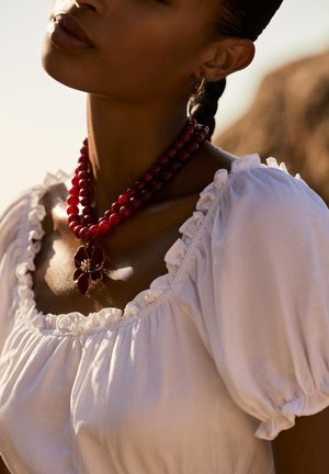 Femme portant un chemisier blanc à volants, un collier de perles rouges à double rang avec un pendentif en fleur, et des boucles d'oreilles en or, sous une douce lumière naturelle.