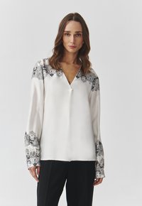 Blusa de satén blanco con escote en V, mangas largas y intrincado bordado floral gris en los puños y el escote. Textura suave, diseño elegante.