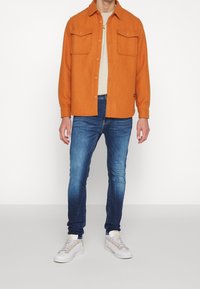Camisa chaqueta de lana naranja con cierre de botón y dos bolsillos en el pecho, combinada con jeans ajustados azules y zapatillas blancas sobre un fondo liso.
