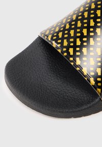 Fekete csúszásgátló papucs fényes fekete és sárga mintás pánttal, amelyen logó található, texturált fekete talppal a tapadásért és kényelemért.