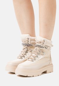 Les bottes en simili cuir beige présentent une semelle épaisse, un col rembourré et des détails en chaîne argentée sur les lacets, offrant un look texturé.