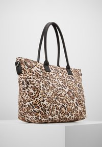 Grand sac fourre-tout en tissu avec imprimé léopard tan, marron et noir, doté de longues anses en cuir noir, posé sur un piédestal blanc.